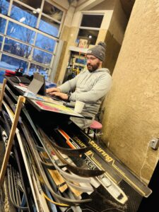 A man with a grey toque and a beard standing in front of a desk that is custom built to hold multiple pairs of skis.