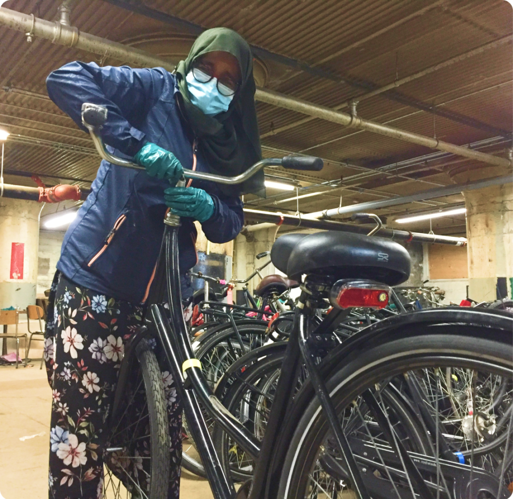 Women wearing PPE, fixing a bike. There are rows of bikes in the background.