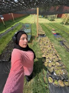 A woman in a pink long sleeve shirt and black headscarf taking a selfie in front of rows of baby trees.
