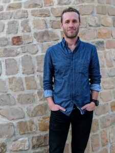 A picture of a white man (dave Haines) in a jean long sleeve button shirt in front of a brick wall