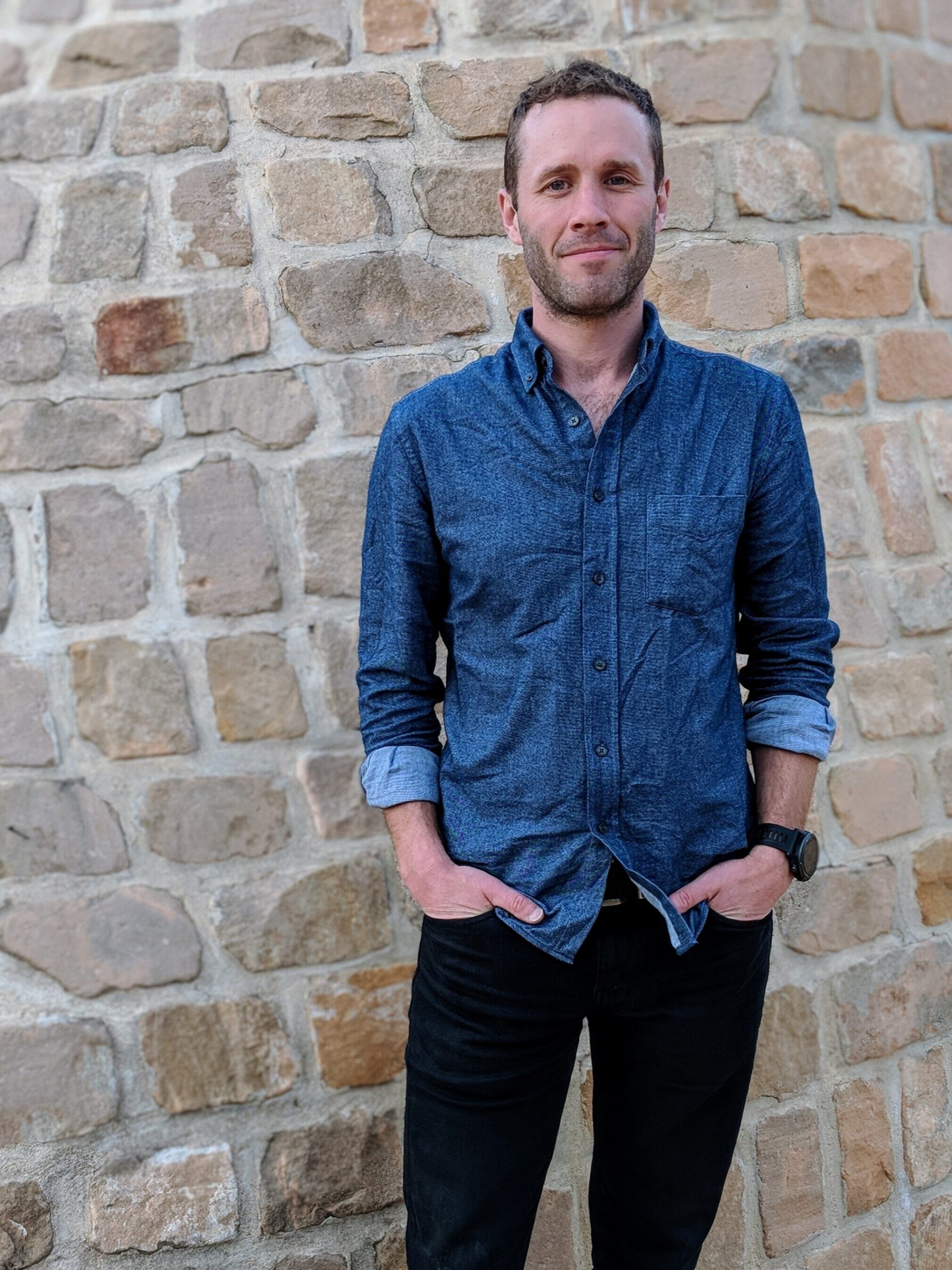 A picture of a white man (dave Haines) in a jean long sleeve button shirt in front of a brick wall